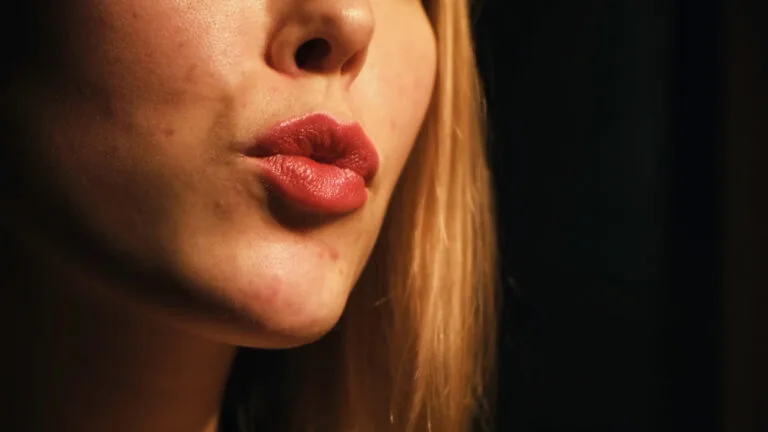 Close-up of woman with blond hair applying the fashion red lipstick on the dark background. Concept natural and volumising lip fillers